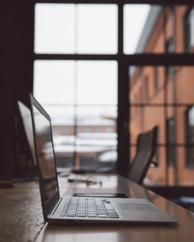 Laptop open on a wooden desk by a large window, overlooking a brick building